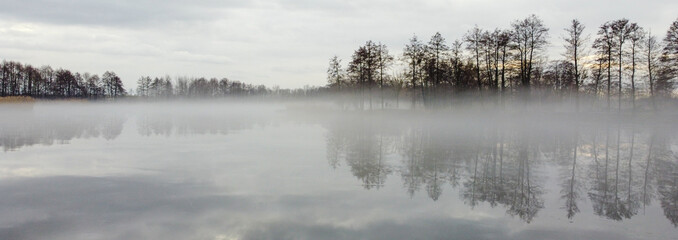 Panoramic view of lake in autumn. Dry trees and yellow grass on the banks. Fog over the lake