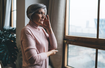 Elderly woman suffering from headache while standing near window