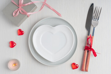 Top view of tableware of round and heart shaped empty plate, metal knife and fork served on white wooden table with glowing candle and gift box prepared for celebration of saint valentine day