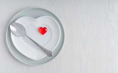 Top view of tableware of round and heart shaped plate with red decoration and metal spoon of silver colour served on white wooden background with copy space for celebration of saint valentine day