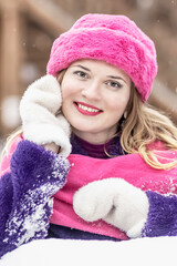 Portrait of a smiling young woman in a bright pink scarf and hat, white mittens and a purple fur coat outdoors in a city park in winter.