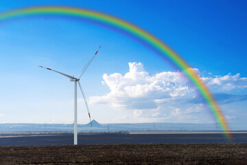 A wind turbine on a flat terrain against a blue sky with a colorful rainbow.
