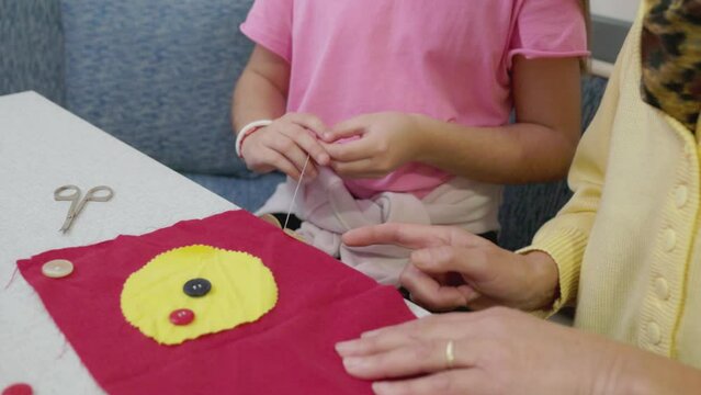 Senior woman teaching her granddaughter, young girl how to sew buttons on a piece of cloth