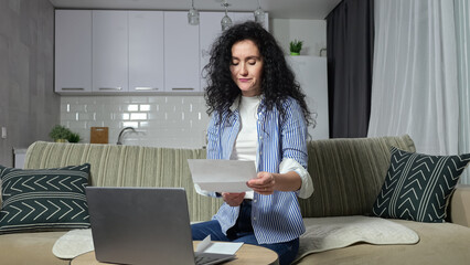 Adult woman checks utility bills and enters information into laptop. Black-haired housewife looks at bills and tries to pay communal payments via Internet from home