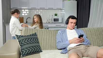 Father wearing headphones listens to music and plays games sitting on sofa at home. Girls siblings...