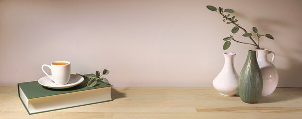 Coffee cup on a gray green book and a group of vases with a sage leaf branch on a wooden table table against a pale wall, panoramic format, home decor, copy space