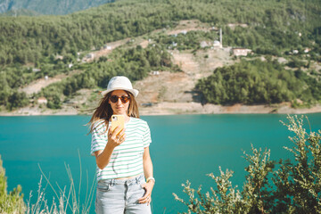 Woman taking selfie on mobile phone of mountains lake background. Traveler female having video chat on cellphone on the blue lake outdoors travel adventure vacation.