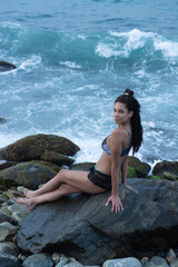 Young woman sitting relaxed looking at the camera with a slight smile, waves and rocks in the background.