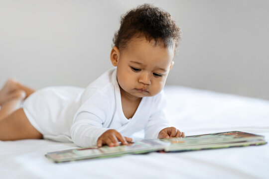 Adorable Black Baby Looking At Pictures In Book While Relaxing In Bed