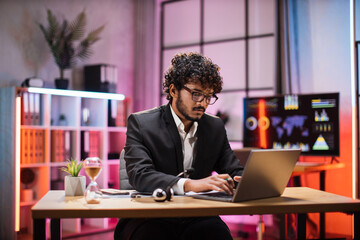 Fototapeta na wymiar Side view, portrait of young smiling experienced smart stylish bearded manager in eyeglass, businessman in suit sitting at table, using laptop in evening modern office