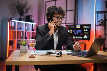 Working process at office of young serious guy having phone call with partners at evening time. Positive bearded businessman having mobile conversation while sitting at table and working on laptop.