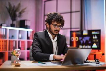 Obraz premium Confident bearded man in formal suit and glasses sitting at desk with modern laptop and looking at screen with serious facial expression. Concept of people and office work.