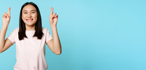 Fototapeta na wymiar Enthusiastic asian girl wishing, cross fingers for good luck and smiling, peeking at camera hopeful, standing over blue background