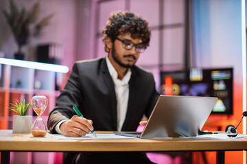 Focus in hand with pen. Attractive confident bearded businessman, office manager, wearing suit using laptop while making financial report while writing on paper working at night.