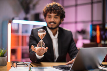 Smiling confident businessman in formal suit sitting at desk with modern laptop and holding hourglass in his hand. Concept of deadline people and office work.