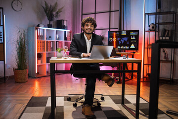 Full length portrait of smiling bearded man in formal suit sitting at desk with modern laptop and looking at camera at evening time at office. Concept of people, deadline and office work.