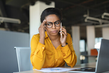 Concerned young black businesswoman talking on cellphone and touching head in stress, having...