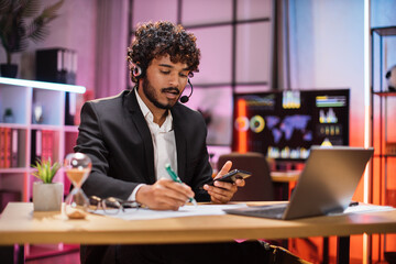 Smiling bearded businessman having video conversation while sitting at table and working on wireless laptop. Working process at office of young guy at evening time.