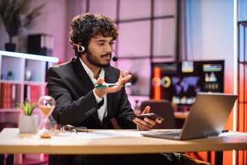 Confident young man at evening time, working process at office. Serious bearded businessman having video conversation while sitting at table and working on wireless laptop.