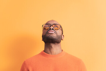 A portrait of a dark-skinned young man looking up in orange sunglasses, the man is wearing a sweater in the same color as the background. Orange color predominates, copyspace.