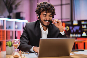 Portrait of confident bearded man in formal wear and headset working at office using laptop. Company worker with beard looking at screen of computer. Concept of work at night, people and business.