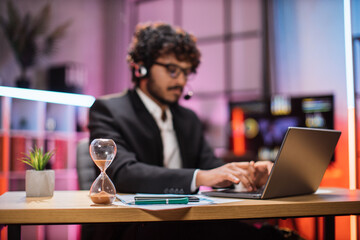 Concept of, overtime, hourglass, deadline, people and office work. Blurred confident businessman in formal suit sitting at desk with modern laptop, focus on holding sand clock in table.