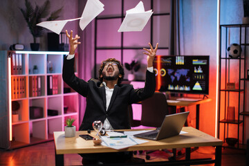 Overjoyed young bearded businessman in suit getting good news, making good deal, throwing papers in air, celebrating and having fun, overtime work at evening office.
