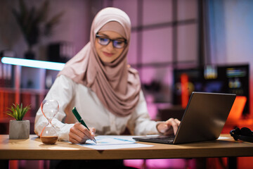 Fototapeta na wymiar Focus in hand with pen. Attractive confident muslim business woman, office manager, wearing hijab using laptop while making financial report while writing on paper working at night.