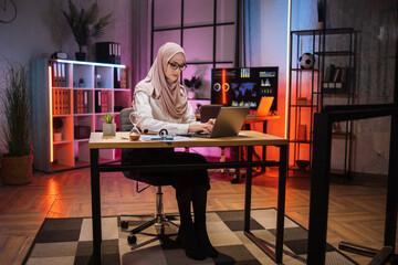 Portrait of attractive confident muslim woman IT specialist, office manager, wearing hijab using laptop making financial report while writing on paper, looking at camera working at night.