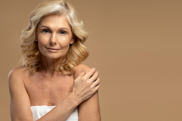 Naklejka premium Close-up beautiful mature woman wrapped in white towel, confidently looking at camera isolated beige background. Copy advertising space. Beauty portrait of 50-55 years senior adult