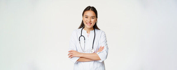 Healthcare and medical concept. Smiling female asian doctor in uniform, looking confident at camera, treating patients, standing over white background