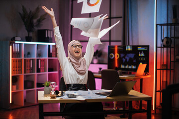 Overjoyed young muslim businesswoman in hijab getting good news, making good deal, throwing papers...