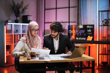 Two multicultural company workers male financial expert and muslim female office worker using laptop, showing paper report with graphs and charts during video meeting with colleagues in office.
