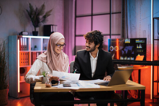 Two Multicultural Company Workers Male Financial Expert And Muslim Female Office Worker Using Laptop, Showing Paper Report With Graphs And Charts During Video Meeting With Colleagues In Office.