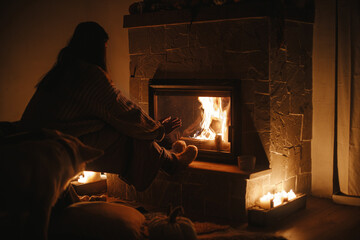 Stylish woman warming up hands and feet at cozy fireplace in evening. Fireplace heating in house,...