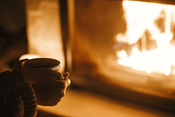 Woman holding cup of tea and warming up hands at cozy fireplace in dark evening room, close up....
