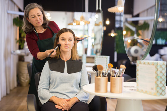 Skilled Elderly Woman Makeup Artist Working In Beauty Studio With Young Female Client, Applying Tonal Foundation On Face.