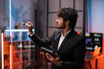 Close up through the glass view of focused young bearded businessman drawing pie chart on glass board , while working in modern office at night time.