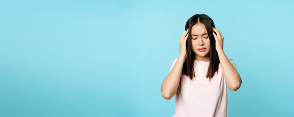 Fototapeta na wymiar Asian young girl with headache, massaging her head with troubled face expression, painful migraine, standing over blue background