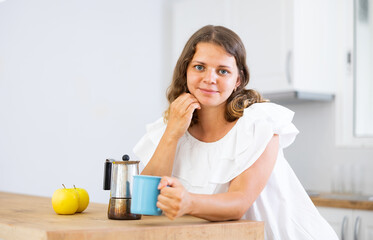 Portrait of happy young woman in white dress drinking morning coffee in modern kitchen