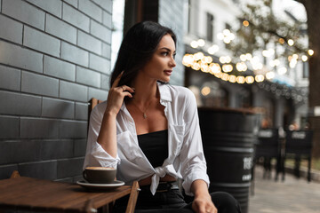 Beautiful successful business woman model in fashion casual clothes with a white shirt sits in a urban cafe on the street and drinks coffee