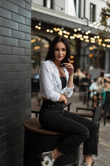 Obraz na płótnie Canvas Fashion beautiful girl in stylish casual clothes with a white shirt and black jeans sits on a barrel near a black brick wall in the city