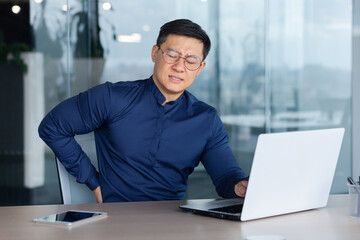 A young male Asian student sits in the office at a desk with a laptop. He feels severe pain in his back, grimaces from pain and fatigue.