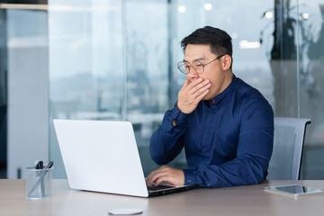 Tired young asian businessman yawns at workplace. He is sitting sleepily at the desk in the office,...