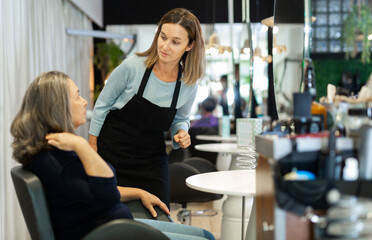 Fototapeta na wymiar Portrait of young woman professional hair stylist talking to elderly female client in salon, choosing new hairdo .