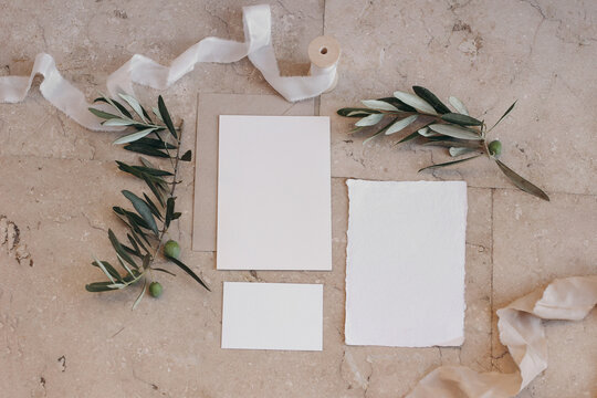 Summer wedding stationery set, suite. Italian, Greek mock-up scene with blank paper greeting cards, envelope on beige marble tiles background. Green olive tree branches, silk ribbon. Flat lay, top.