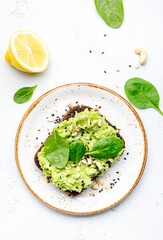 Avocado sandwich or toast on rye bread with spinach, crushed cashew nuts and sesame seeds, on plate, white table background, top view
