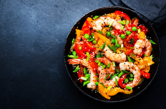 Stir fry with shrimps, red and yellow paprika, green pea, chives and sesame seeds in frying pan. Asian cuisine dish. Black stone kitchen table background, top view