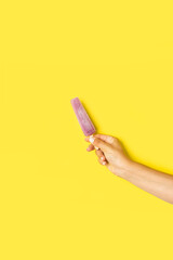 Mano de mujer sosteniendo una paleta de helado natural de sabor a uva sobre un fondo amarillo...