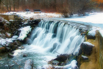 Fototapeta premium On a Winter day in Wisconsin, a slow shutter speed smooths a small waterfall in a forest with two nearby benches.